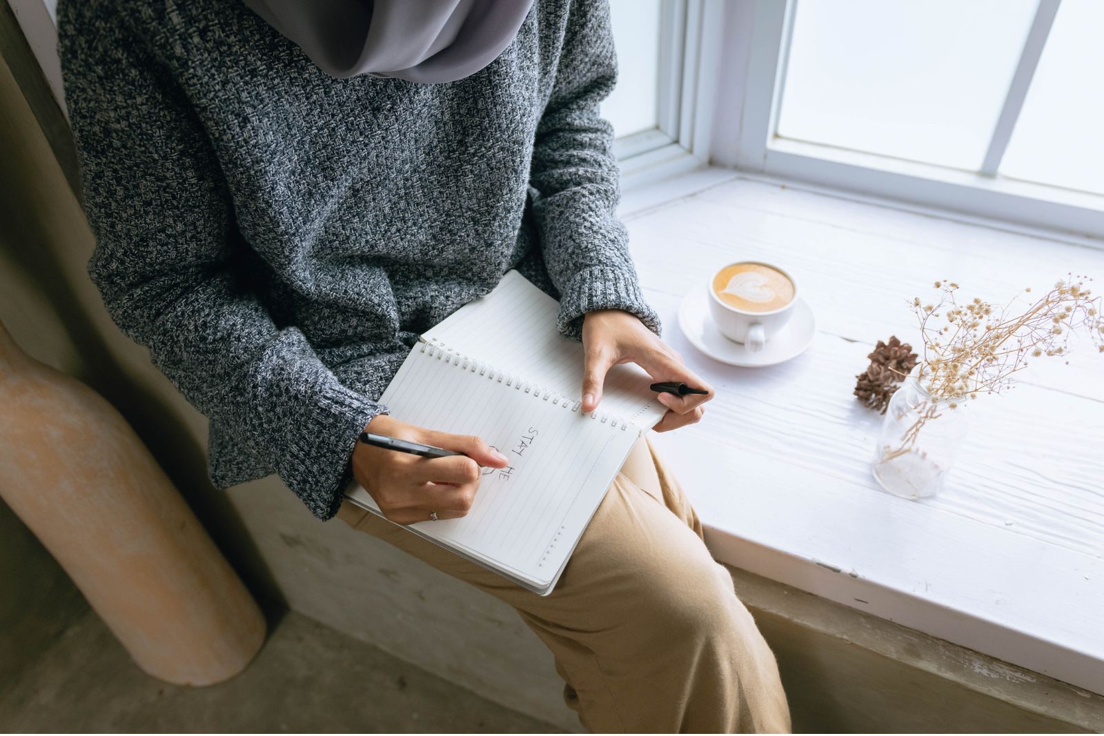 Adult writing in a notebook at a desk near a window